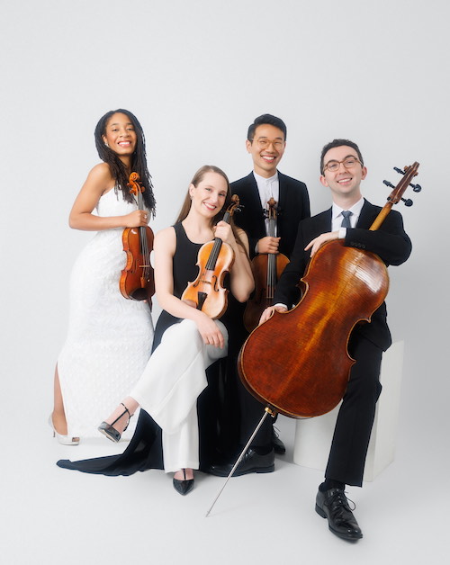 Four musicians wearing black and white, smile while posing with their instruments: two violins, a viola, and cello.