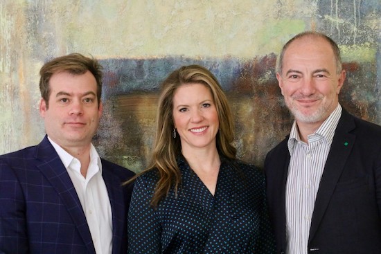 A smiling woman between two smiling men indoors, with a large abstract painting behind.