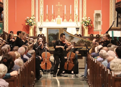 A violinist performs a solo with two cellos, a harpsichord, and other string musicians behind him in a church with audience visible in pews.